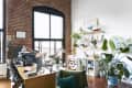 Loft office with brick walls, large windows, wooden desk, plants, and shelves with decor and books.