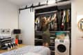Woman organizing clothes in a bedroom closet with sliding doors, next to a bed with patterned pillows and a washing machine.