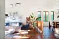 Living room with beige sofa, blue chair, gallery wall, large plant, and dining area with wooden table and chairs.