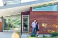 Couple standing outside a modern house with large windows, wood siding, and decorative plants.