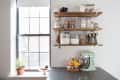 Kitchen counter with mint green mixer, fruit basket, and wooden shelves holding jars of ingredients.