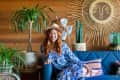 Woman in blue floral dress and hat sitting on blue sofa, surrounded by plants and sunburst wall decor.