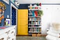 Colorful children's room with a yellow door, blue walls, white shelves filled with books, toys, and a white chair.