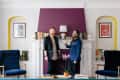 Two men standing by a white fireplace with books, purple wall, and orchid in a stylish living room.