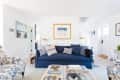 Coastal-themed living room with blue sofa, striped pillows, floral armchairs, and nautical artwork on white shiplap walls.