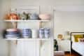 Open shelving with teapots, bowls, and plates in a kitchen, adjacent to a bedroom with colorful pillows.