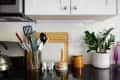 Kitchen counter with utensil holder, cutting boards, potted plant, kettle, and colorful jars against a tiled backsplash.