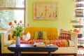Cozy living room with a cream sofa, colorful pillows, yellow throw, floral vase, and books on a floating shelf.
