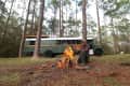 Man sitting by a campfire in a forest with a converted green bus in the background.