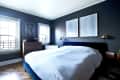 Bedroom with dark blue walls, white bedding, wooden dresser, two framed artworks, and brass wall lamps.