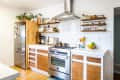 Modern kitchen with stainless steel appliances, wooden cabinets, open shelves with jars, and a bowl of fruit on the counter.