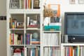 Bookshelf with colorful books, magazines, vases, framed art, and a TV on a wooden floor with a patterned rug.