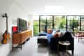 Living room with blue sectional sofa, colorful cushions, wall-mounted guitar, wooden sideboard, and large garden-view windows.