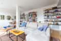 Spacious living room with beige sectional sofa, colorful pillows, bookshelves, and a small dining area with round mirror.