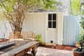 Patio with wooden table, rattan bench, potted plants, and a white door under a tree.