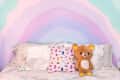 Bed with pastel rainbow wall, pink pillow with eye and hand prints, and a brown teddy bear.
