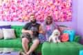 Couple sitting on a green sofa with two dogs, in front of a vibrant pink floral wall.