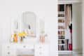 White vanity with mirror, yellow flowers, perfumes, and makeup brushes beside an open closet with shoes and hanging clothes.