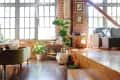 Loft living room with brick walls, large windows, green plants, a gray sofa, and a wooden floor with steps.