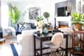 Living room with black sofa, white armchair, fireplace, plants, and a black desk with books and flowers.