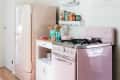 Retro kitchen with pink fridge and stove, white cabinet, wooden utensils, and vintage tins on a shelf.