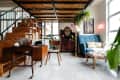 Loft-style room with wooden stairs, desk with laptop, blue sofa, plants, and vintage dresser under string lights.