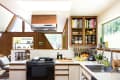 Mid-century modern kitchen with wooden cabinets, open shelving, jars, cookbooks, and a fruit bowl near a large window.