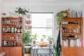 Home bar with wooden shelves, liquor bottles, glassware, plants, books, and a small table with oranges by a window.