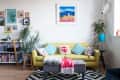 Yellow sofa with colorful cushions, surrounded by plants, art, and bookshelves in a cozy living room.