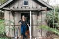 Woman standing in the doorway of a small brown cabin surrounded by greenery.