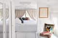 Cozy bedroom with white bedding, colorful pillows, framed art, and books on a shelf, featuring a window with beige curtains.