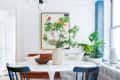 Dining area with round marble table, black chairs, large plant centerpiece, abstract wall art, and potted plants by window.
