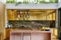 Modern kitchen with gold cabinets, marble backsplash, pink island, and plants above. Two stools and fruit bowl on the counter.