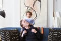 Man sitting on a blue sofa with a child on his shoulders, abstract art in the background, and patterned cushions.