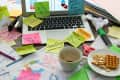 Cluttered desk with sticky notes, laptop, coffee cup, waffle, and crackers, indicating a busy work environment.