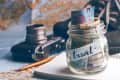 Jar labeled "travel" filled with various currencies, next to a vintage camera, notebook, and pencil on a map.