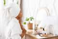 Woman in a white robe and towel looking in a mirror on a wooden vanity with makeup items and a potted plant.