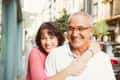 Woman in pink sweater hugging smiling older man on a sunny city street.