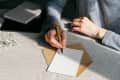 Person writing on a blank card with a pen, next to an open envelope and a small notebook on a table.