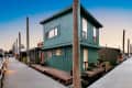 Two-story green houseboat with large windows and potted plants on a wooden dock at sunset.