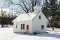 Small white cottage with a snow-covered roof, pink door, and yellow patio set in a snowy landscape.
