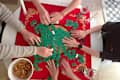 Group of hands playing a tile game on a red tablecloth, surrounded by snacks and candles.