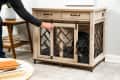 Person closing a wooden dog crate with a black dog inside, next to a table with stacked books.