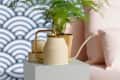Cream watering can and gold plant pot with fern on white pedestal, against a patterned wall.