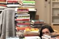 Stacks of colorful books piled high in a room, with a person peeking through the stacks.