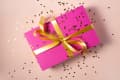 A bright pink box with a gold ribbon on it and heart confetti on top