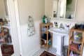Bathroom with pedestal sink, wooden shelves, toiletries, and a mirror. Green towel on a ring holder and a potted plant visible.