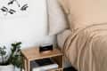 Bedside table with digital clock, potted plant, books, and beige bedding in a minimalist bedroom.