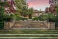 Stone steps leading to a large house with lush greenery, trees, and planters at sunset.