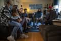 Group of six people sitting in a living room, one in a wheelchair, with blue abstract art on the wall.